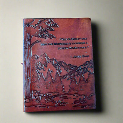 Brown leather-bound book with mountain and tree design and John Muir quote on a beige background