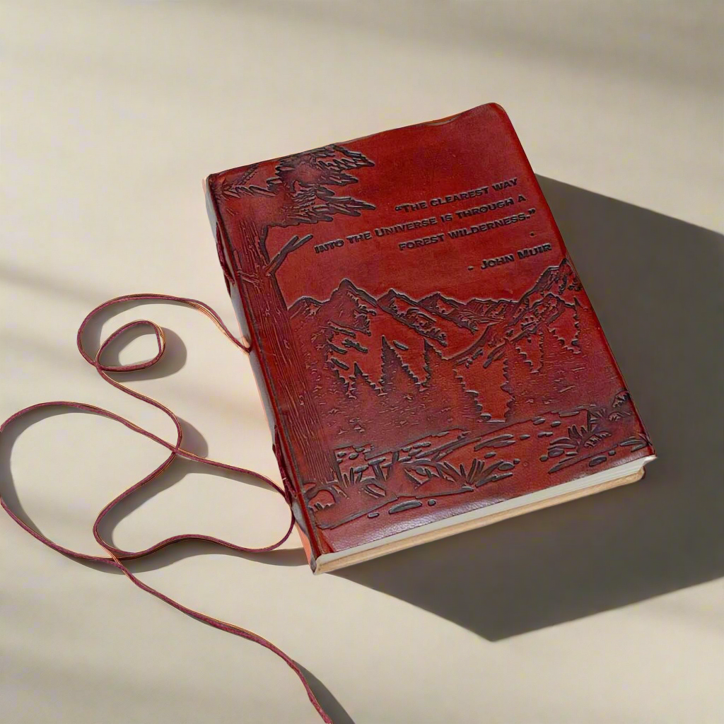 A red leather journal with embossed mountain and forest designs and the quote by John Muir 'The clearest way into the Universe is through a forest wilderness.' It has a string tie closure and is placed on a white wooden surface.