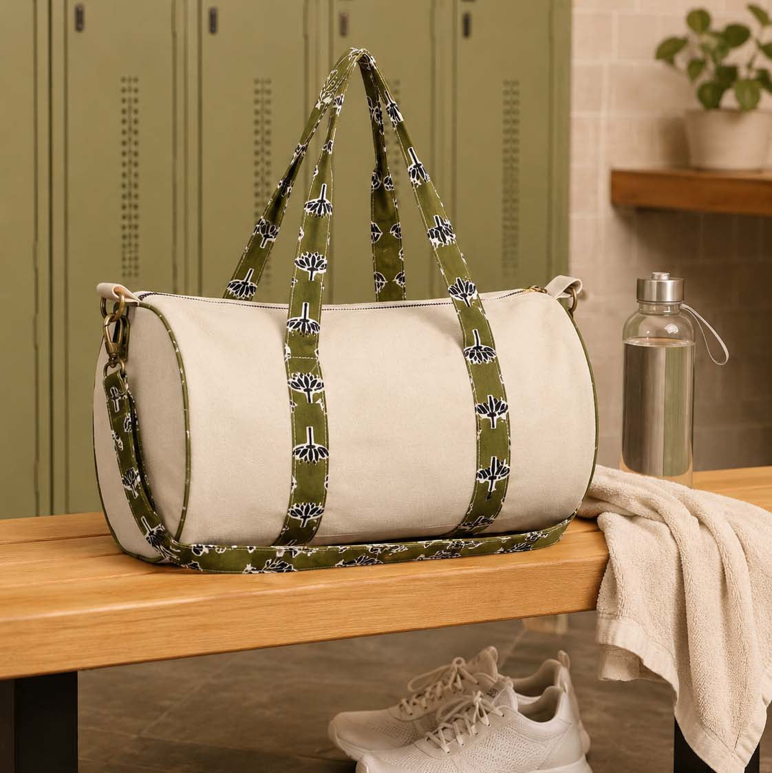 A cylindrical duffel bag with off-white panels and olive green patterned straps sits on a wooden bench in a locker room setting, with sneakers and a towel nearby.