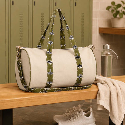 A cylindrical duffel bag with off-white panels and olive green patterned straps sits on a wooden bench in a locker room setting, with sneakers and a towel nearby.