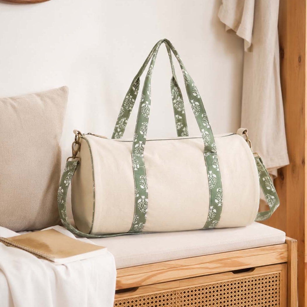 A beige canvas duffel bag with green floral patterned shoulder straps and side panels, resting on a wooden bench in a softly styled room.