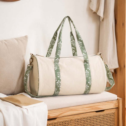 A beige canvas duffel bag with green floral patterned shoulder straps and side panels, resting on a wooden bench in a softly styled room.